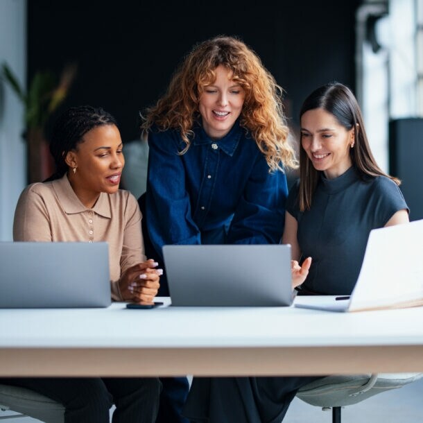 Drei Frauen schauen gemeinsam auf ein Laptop auf einem großen Tisch mit zwei weiteren Laptops in einem modernen Büro.