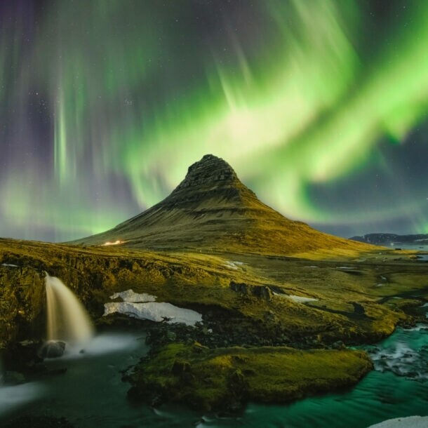 Moosbedeckte Vulkanlandschaft mit Wasserfällen unter Nachthimmel mit grün und lila leuchtenden Polarlichtern.