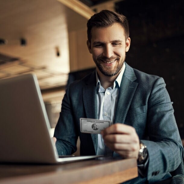 Ein lächelnder Geschäftsmann sitzt an einem Tisch vor einem Laptop und blickt auf eine silberne Platinum Kreditkarte von American Express in seiner Hand.