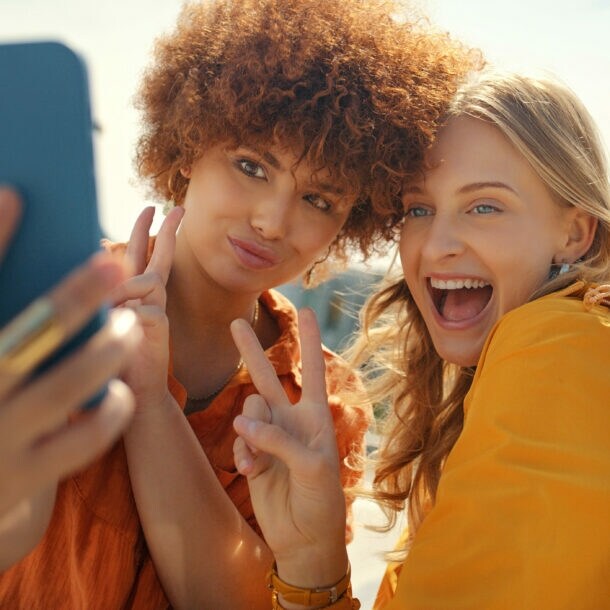 Zwei junge Frauen zeigen Peace-Zeichen mit ihren Fingen, während sie ein Selfie per Smartphone aufnehmen unter freiem Himmel.
