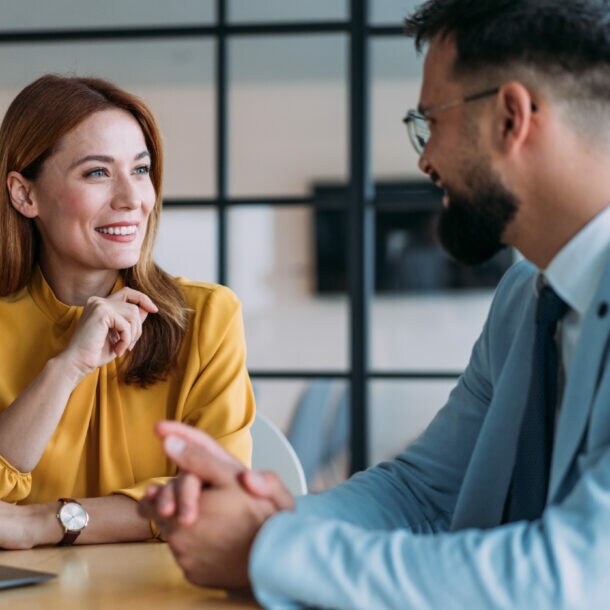 Eine Frau und ein Mann in eleganter Kleidung führen ein freundliches Gespräch an einem Tisch in einem Büro.