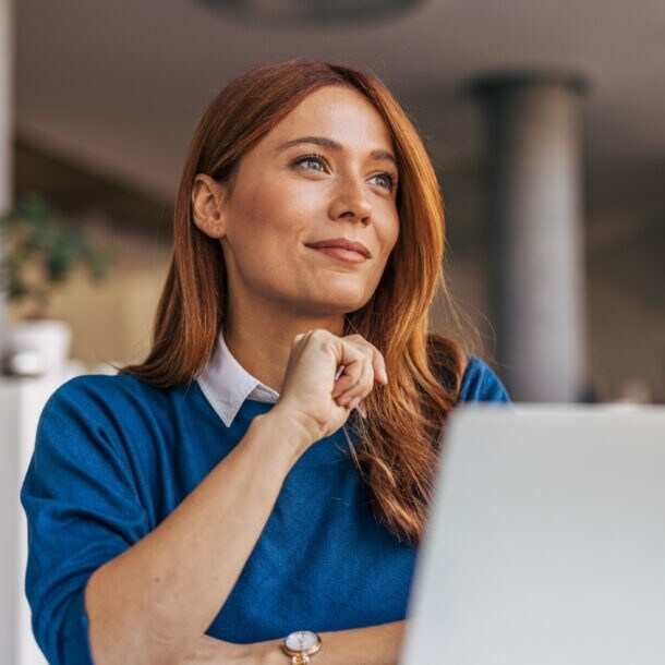 Eine Frau im blauen Pullover sitzt vor einem Laptop und schaut zufrieden lächelnd in ein Büro.