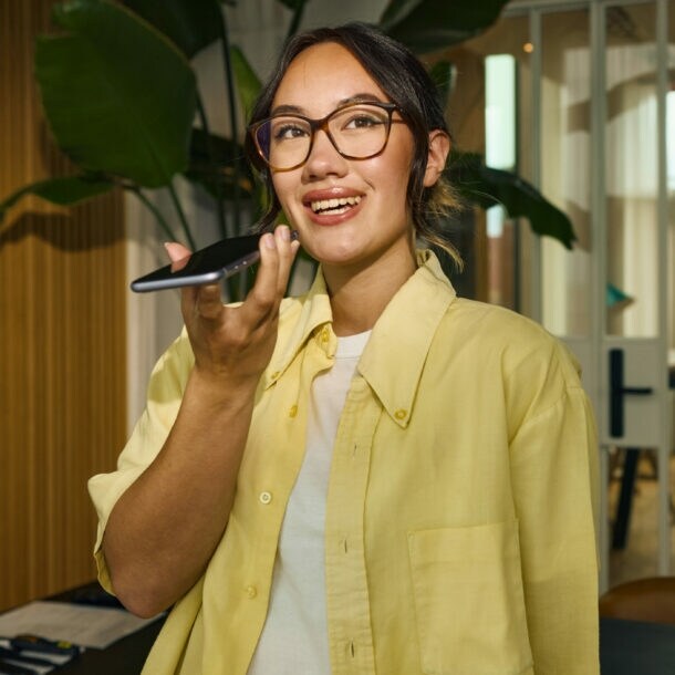 Eine junge Frau im gelben Hemd spricht in ein Smartphone in einem Büro.