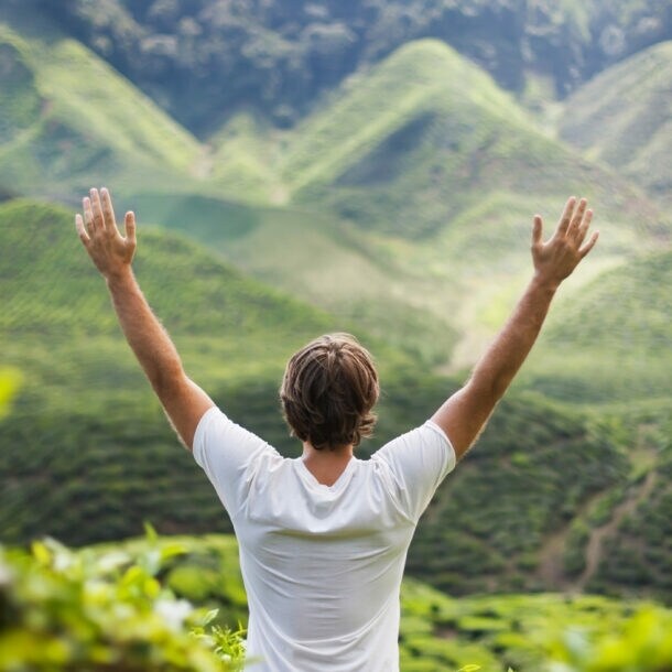 Mann mit weißen T-Shirt steht mit erhobenen Armen vor grünen, bewaldeten Hügeln.