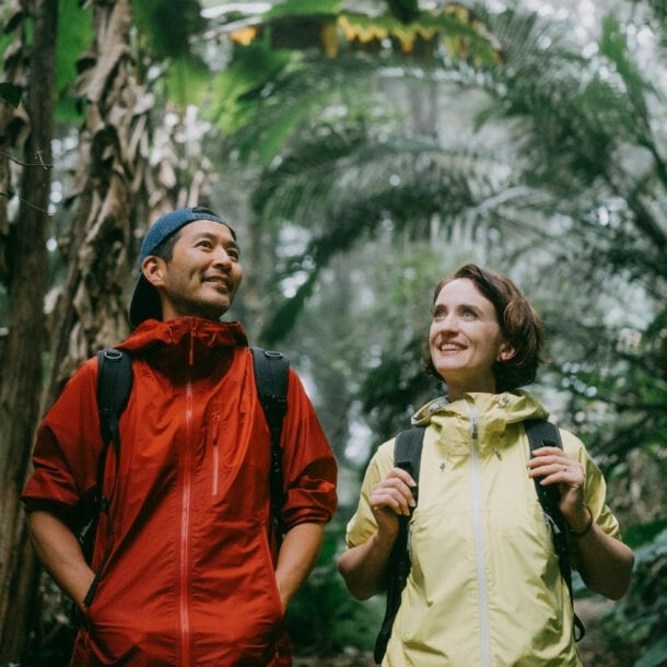 Zwei Wandernde mit Rucksäcken in einem dichten Dschungel, einer trägt eine rote Jacke, die andere eine gelbe Jacke.
