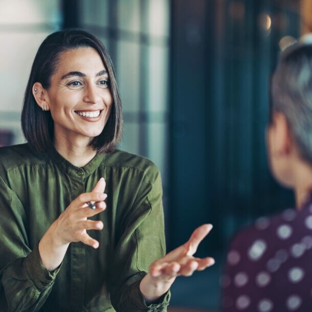 Eine lächelnde Frau in grüner Bluse im Gespräch mit einer nur von hinten erkennbaren Person.