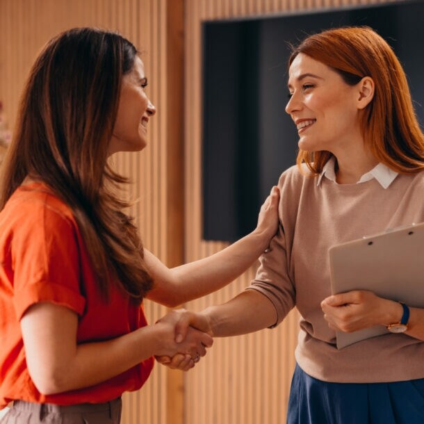 Zwei Frauen geben sich die Hand und lächeln sich an. Eine Frau legt die Hand auf den Arm der anderen Frau.