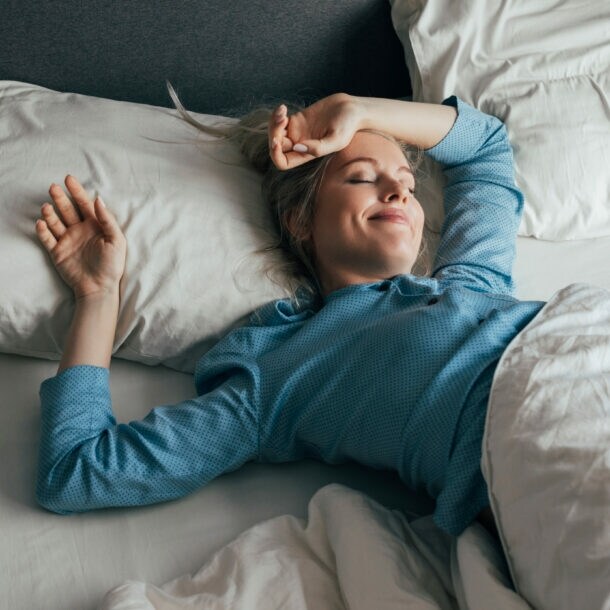 Eine zufrieden lächelnde Frau im blauen Pyjama liegt mit geschlossenen Augen in einem Bett mit weißer Bettwäsche bei Tageslicht.