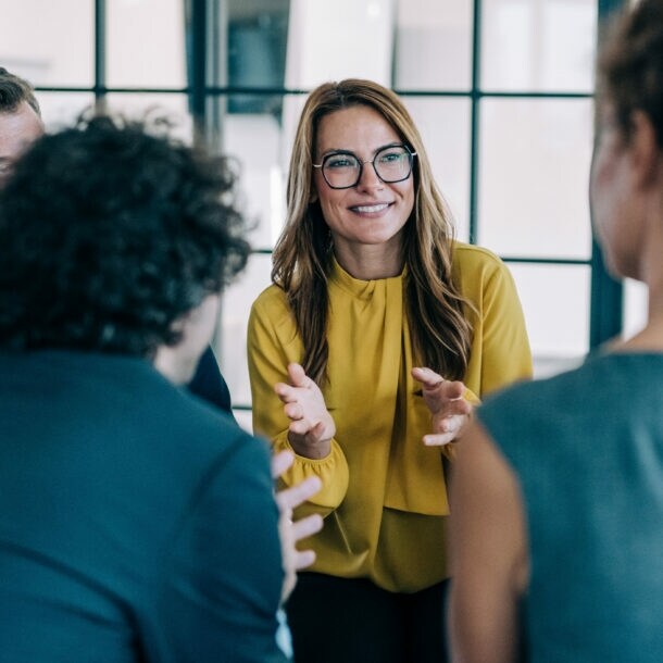Mehrere Personen in Business-Kleidung stehen in einem Kreis, im Zentrum eine lächelnde, gestikulierende Frau im gelben Oberteil.
