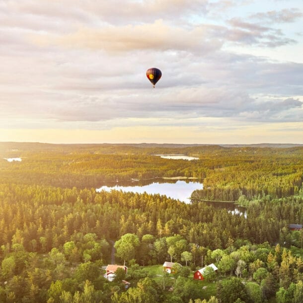 Heißluftballon mit rotem Muster fliegt über bewaldete Landschaft mit mehreren Seen und vereinzelten Häusern bei Sonnenuntergang.