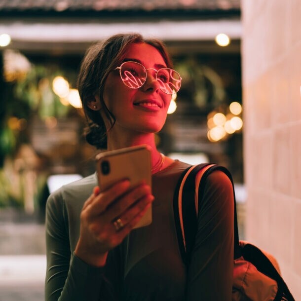 Eine junge Frau hält lächelnd ein Smartphone vor sich in der Hand, in ihrer Brille spiegelt sich der Schriftzug eines roten Logos an einer Wand.