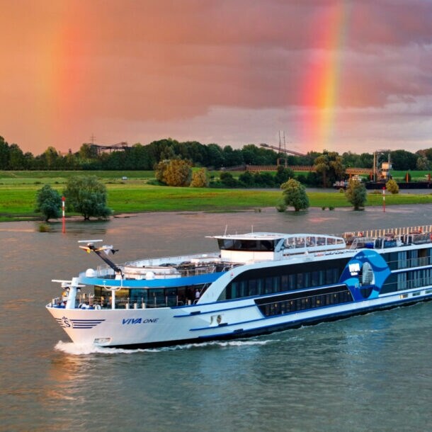 Flusskreuzfahrtschiff Viva One fährt vor grünem Flussufer mit Regenbogen am bewölkten Himmel.