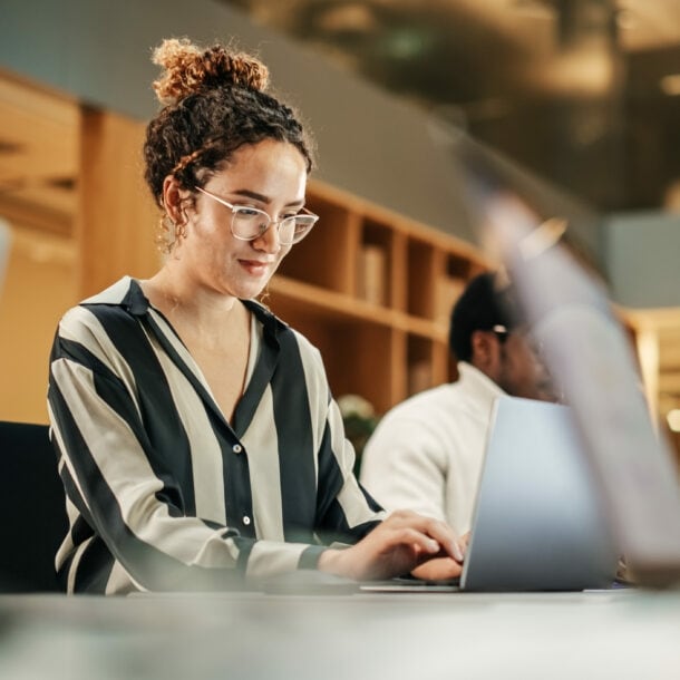 Eine junge Frau in gestreifter Bluse arbeitet an einem Laptop an einem Tisch mit weiteren Personen in einem Büro.