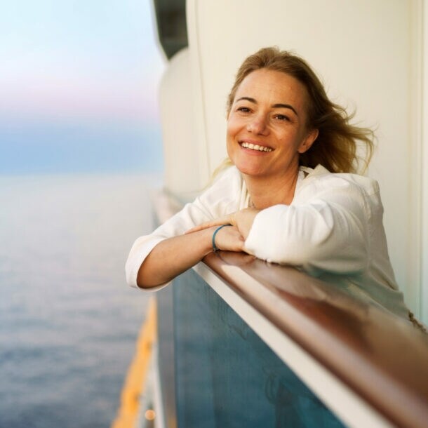 Eine lächelnde Frau lehnt an der Reling einer Balkonkabine auf einem Kreuzfahrtschiff auf See bei Abenddämmerung.