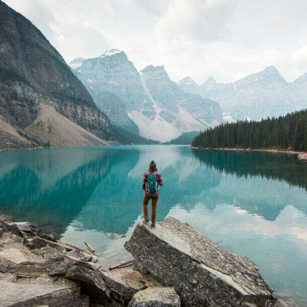 Person mit rotem Rucksack steht auf Felsen am Ufer eines klaren Bergsees, umgeben von Nadelwald und hohen Bergen.