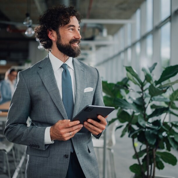 Halbporträt eines lächelnden Mannes im grauen Anzug mit Einstecktuch und Krawatte, der mit einem Tablet in einem Büro steht.