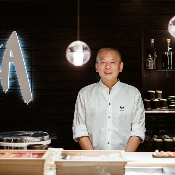 Ein Küchenchef steht hinter einer Sushibar in einem Restaurant mit einem Wandlogo Aska.