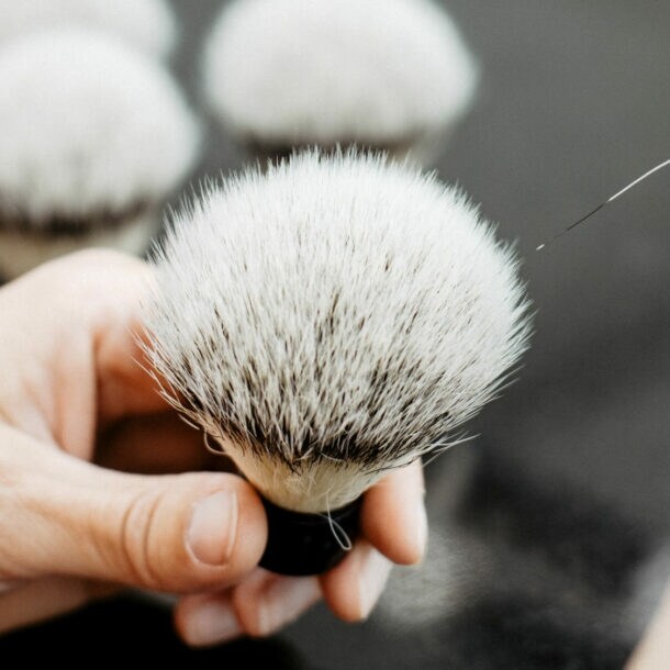 Hand hält einen weißen Rasierpinsel mit dunklen Borsten, eine zweite Hand führt eine Pinzette an die Borstenspitze.
