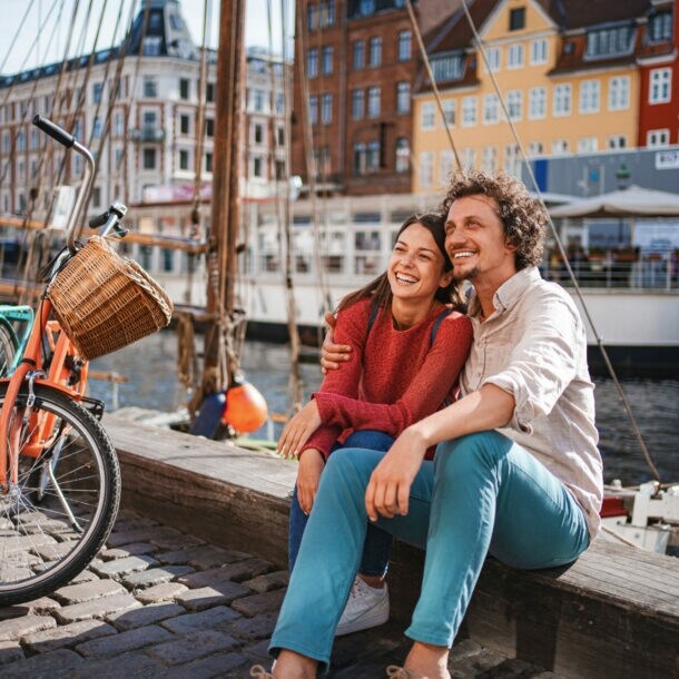 Ein lachendes Paar sitzt eng beieinander auf einer Kaimauer an einem Kanal mit bunten Häusern im Stadtzentrum bei Sonnenschein, neben ihnen stehen zwei Fahrräder auf Kopfsteinpflaster.
