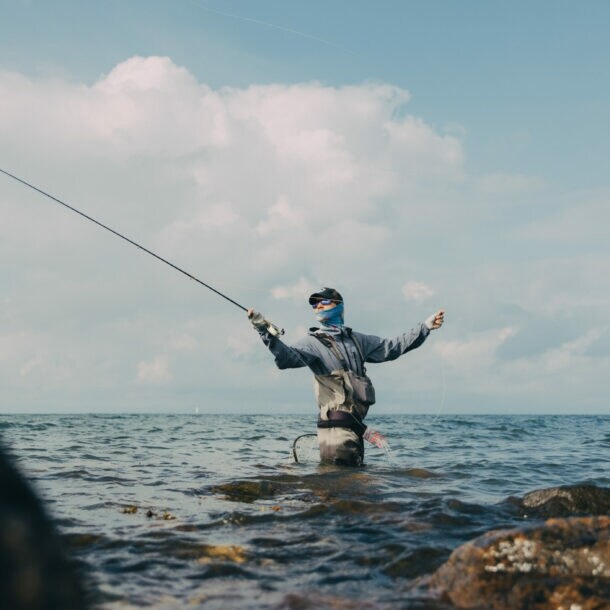 Angler steht hüfttief im Wasser zwischen Felsen und wirft eine Angelrute aus, bewölkter Himmel im Hintergrund.