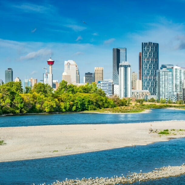 Skyline von Calgary mit Fernsehturm vor blauem Himmel, im Vordergrund Grünflächen und Sandbänke entlang eines flachen Flussufers.