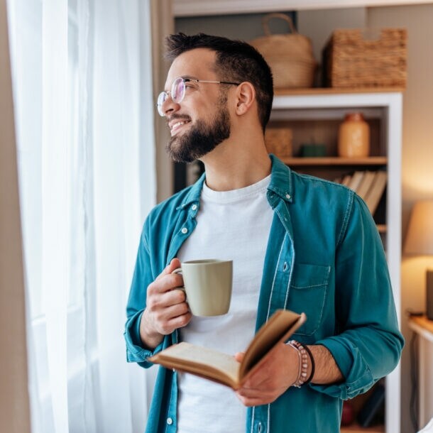 Ein Mann im Jeanshemd mit Buch und Kaffeebecher in den Händen schaut entspannt aus einem Fenster mit Vorhängen.