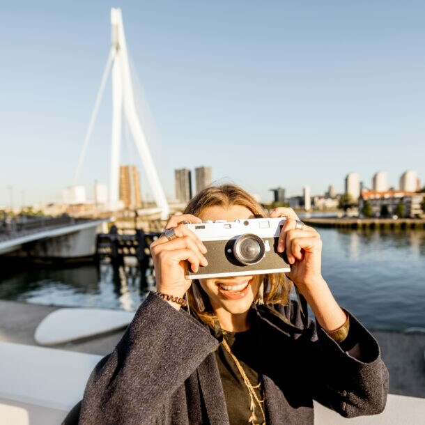 erson mit dunkelgrauem Mantel hält eine weiße Sofortbildkamera vor das Gesicht, im Hintergrund die Skyline von Rotterdam und eine Brücke über Wasser.