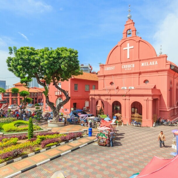 Belebter Platz mit Parkanlage, umgeben von pinkfarbenen Kolonialbauten samt Kirche unter blauem Himmel.