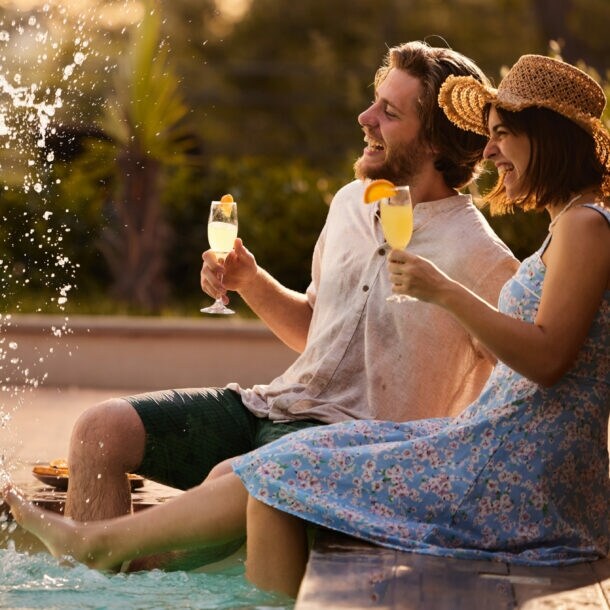 Eine Frau und ein Mann sitzen am Rand eines Pools mit einem Getränk in der Hand und spritzen mit ihren Füßen.