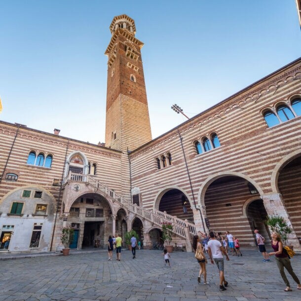 Personen in einem italienischen Palastinnenhof mit gestreifter Fassade und Außentreppe unter einem Turm vor blauem Himmel.