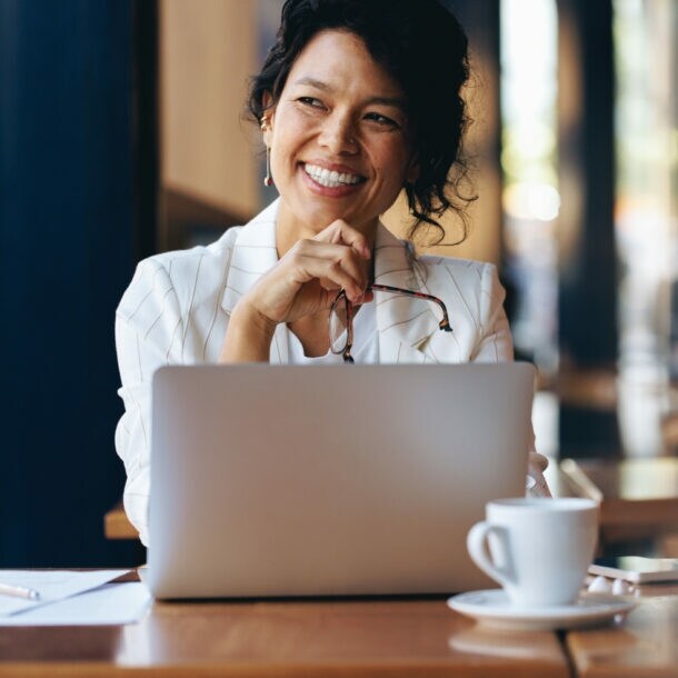 Eine Frau sitzt zufrieden lächelnd an einem Laptop an einem Tisch mit Kaffeetasse in einem Café.