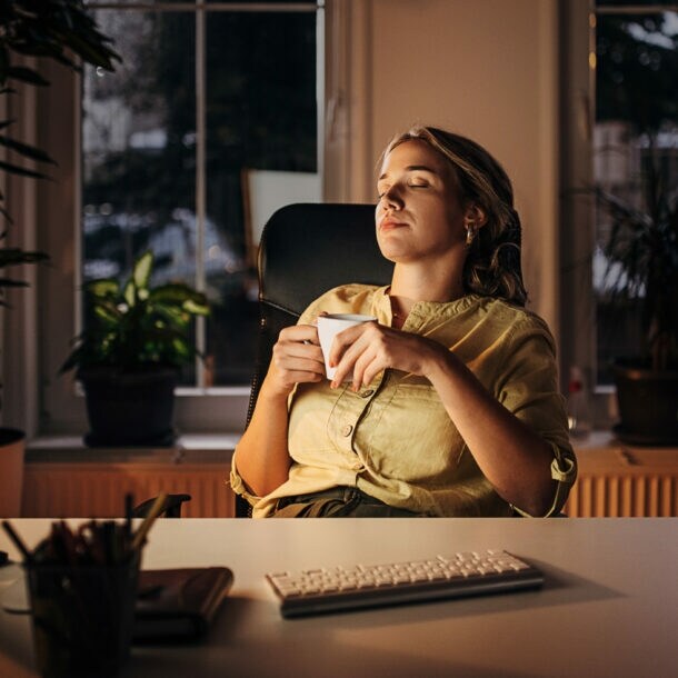 Eine Frau mit einer Tasse in den Händen lehnt mit geschlossenen Augen in einem Schreibtischstuhl an einem Schreibtisch am Abend.
