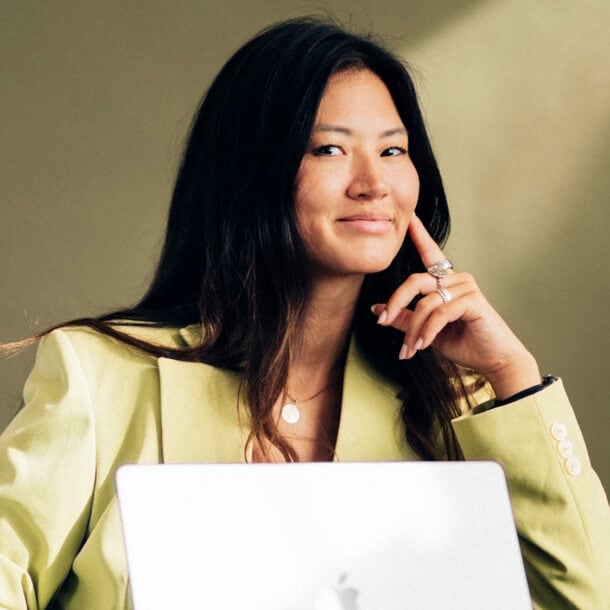 Eine Frau in einem hellgelben Blazer mit aufgestütztem Arm und einem aufgeklapptem Laptop in einem Sessel lächelt dezent in die Kamera.