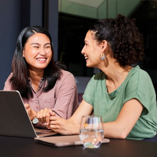 Zwei sich zugewandte lachende Frauen sitzen nebeneinander an einem Tisch, vor ihnen steht ein Laptop.
