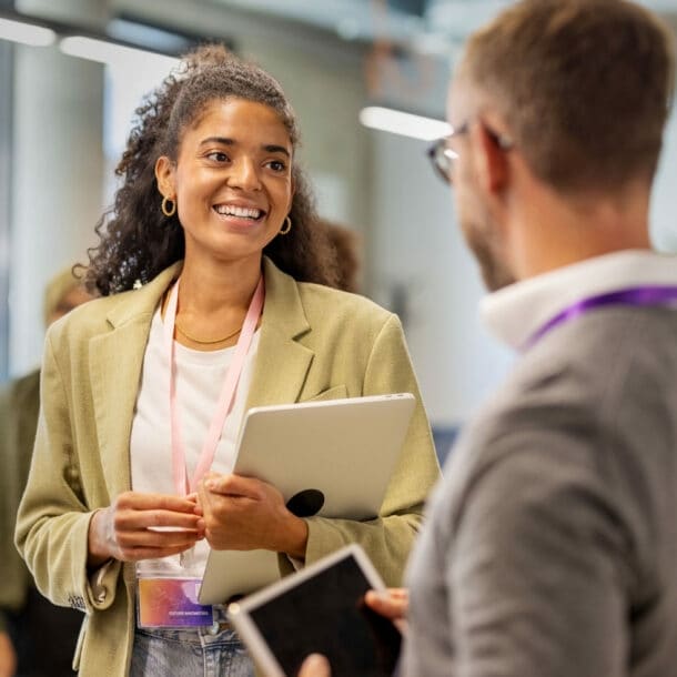 Eine junge Frau im Blazer mit Laptop in der Hand steht in einem Konferenzraum und unterhält sich mit zwei weiteren Personen.