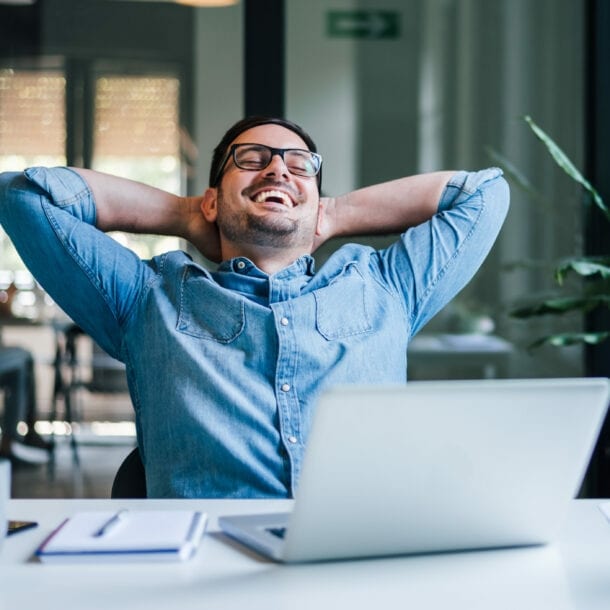 Ein lachender Mann lehnt entspannt zurück mit verschränkten Armen im Nacken auf einem Stuhl an einem Schreibtisch.
