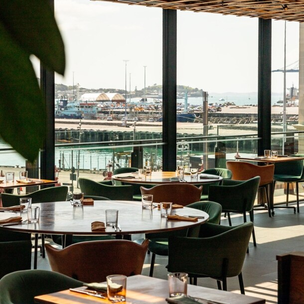 Runde Esstische in einem Restaurant mit Blick durch Panoramafenster auf ein Hafengebiet.