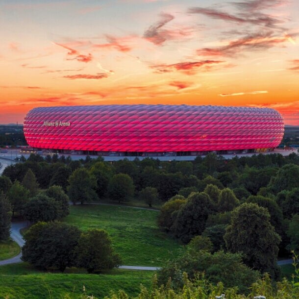 Panoramaaufnahme eines pink leuchtenden Fußballstadions unter orange gefärbtem Himmel bei Sonnenuntergang, im Vordergrund Wiesenfläche mit Wald und Kapelle.