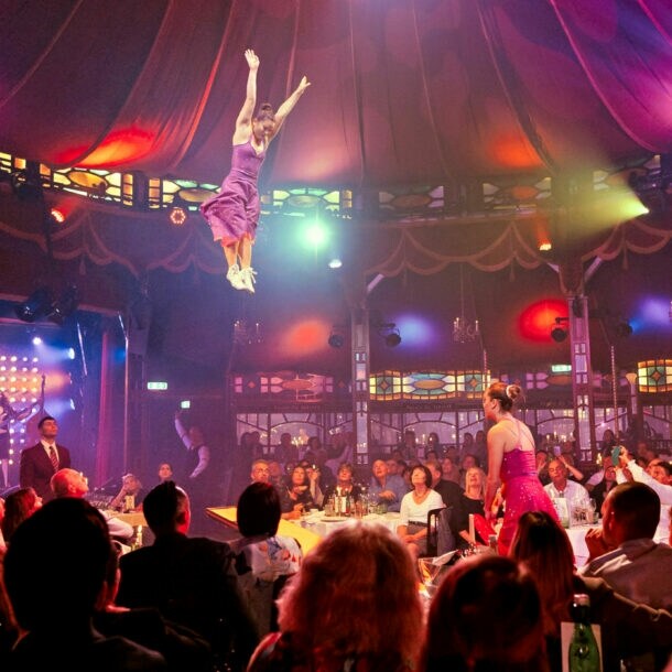 Personen sitzen beim Dinner an Tischen in einem Spiegelzelt während einer Show mit Akrobatinnen.