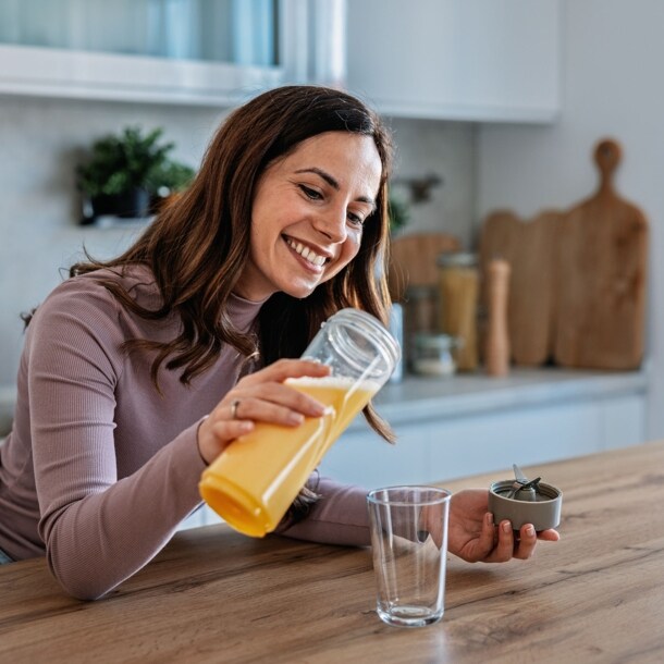 Eine lächelnde Frau gießt ein gelbes Getränk in eine Glas an einer Küchentheke.