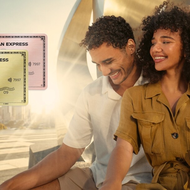 Ein lächelndes Paar sitzt vor einer gold glänzenden Metallskulptur im Sonnenschein auf einem weitläufigen Platz, er hat einen Arm um sie gelegt.