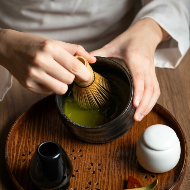 Nahaufnahme von zwei Händen bei der Zubereitung eines grünen Tees mit einem Bambusbesen in einer dunklen Keramikschale auf einem Holztisch.