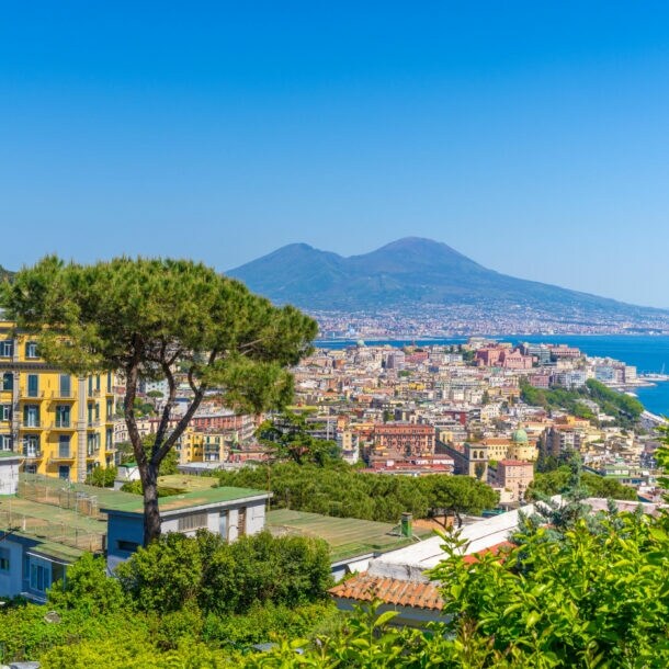 Blick von oben auf die Häuser von Neapel, im Hintergrund Meer und ein Vulkan.