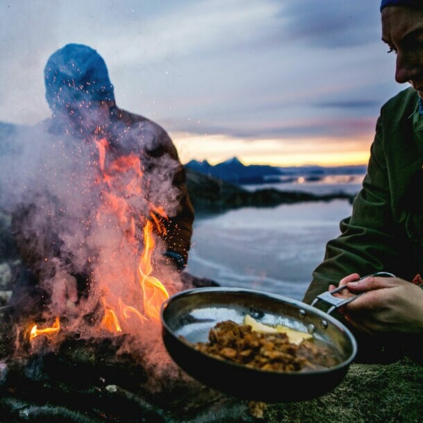 Zwei Personen bereiten Speisen in Pfannen über einem Feuer an einer schroffen Felsküste zu bei Abenddämmerung.