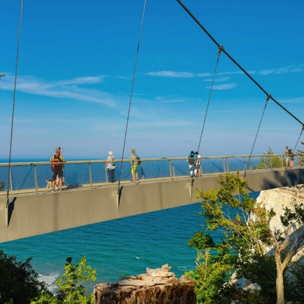 Mehrere Personen laufen auf einer schmalen Brücke zwischen Kreidefelsen am Meer unter blauem Himmel bei Sonnenschein.