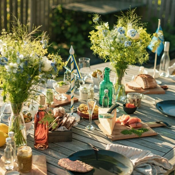 Gedeckter Holztisch für ein Mittsommer-Essen im Freien, mit Brot, gebeiztem Lachs, Käse, Eingelegtem, Getränken und frischen Blumensträußen.