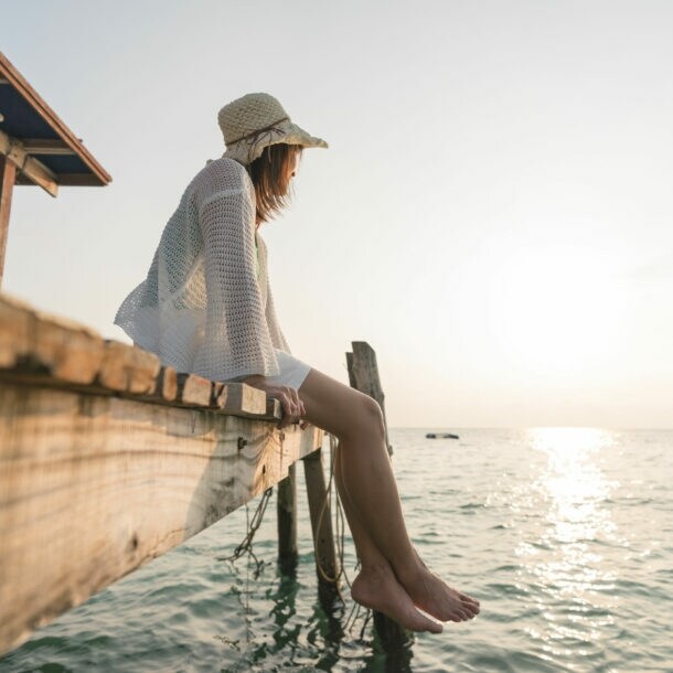 Frau mit Sonnenhut und barfuß, die auf einem Holzsteg sitzt und aufs Wasser schaut.