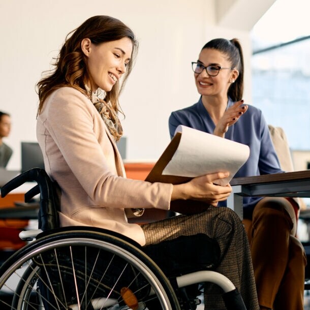 Eine Frau im Rollstuhl mit Klemmbrett in der Hand sitzt an einem Tisch neben einer weiteren Frau in einem hellen Raum.