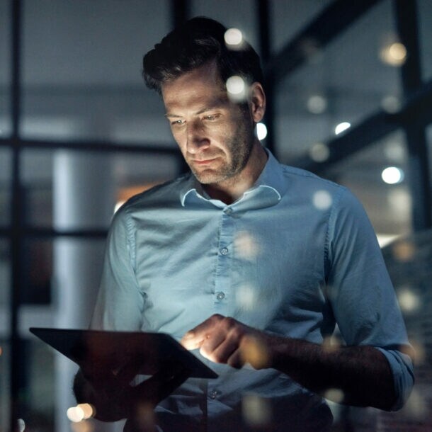 Ein Mann steht in einem dunklen Raum und hält ein Tablet in seinen Händen, dessen Displaylicht sein Gesicht anleuchtet.