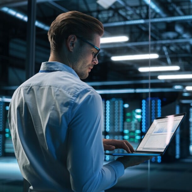 Profil eines Mannes im Hemd, der mit einem aufgeklappten Laptop in einem Rechenzentrum steht.